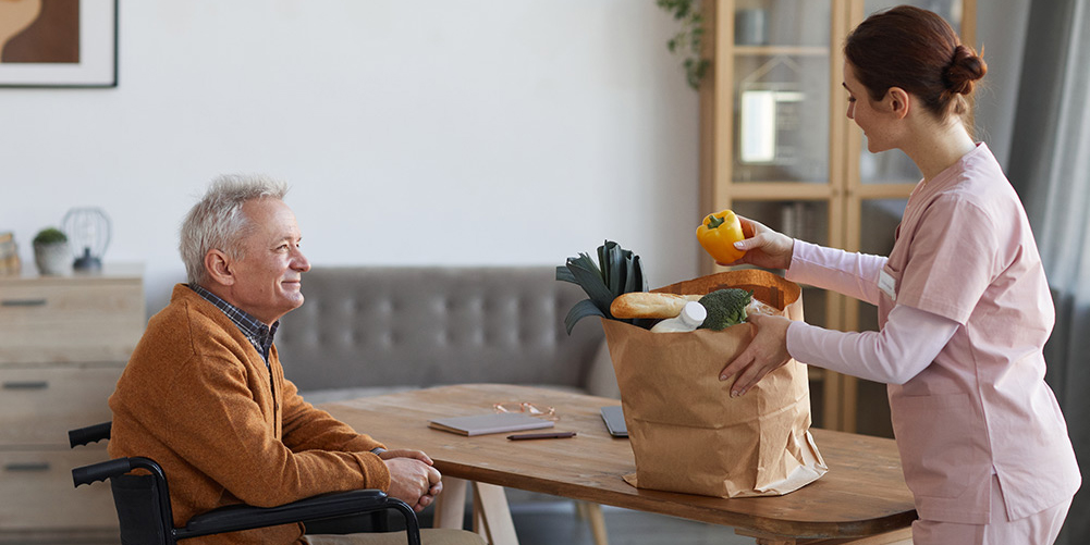 Care worker woman unpacking groceries for senior man in wheelchair